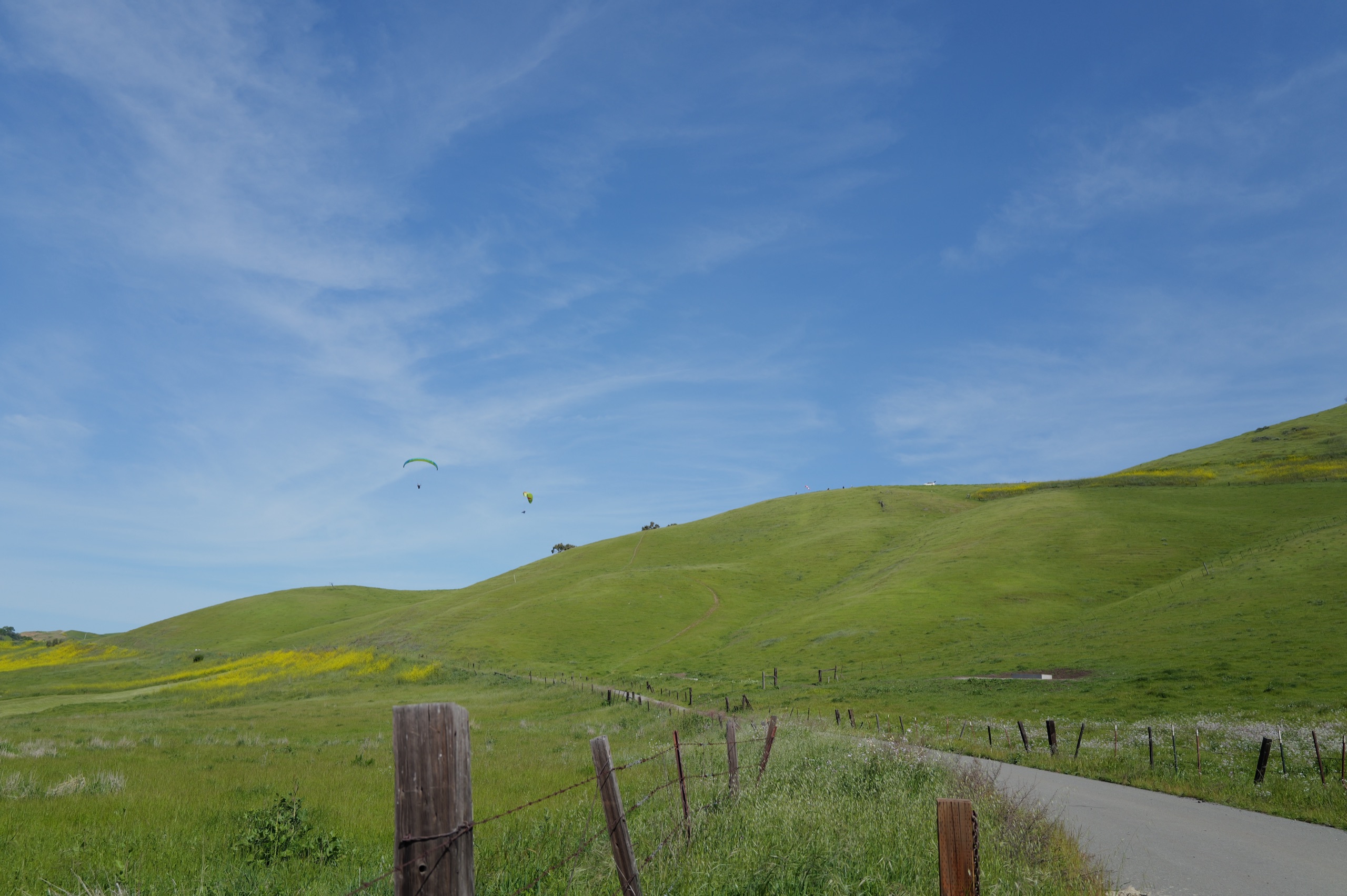 Two paragliders flying over rolling green and yellow hills, fence in the foreground, blue sky above
